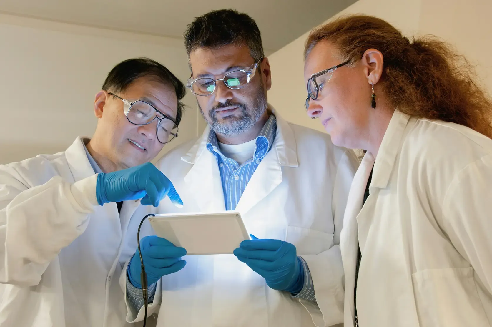 Three analysts in laboratory coats examining a backlit document together, one pointing at it.