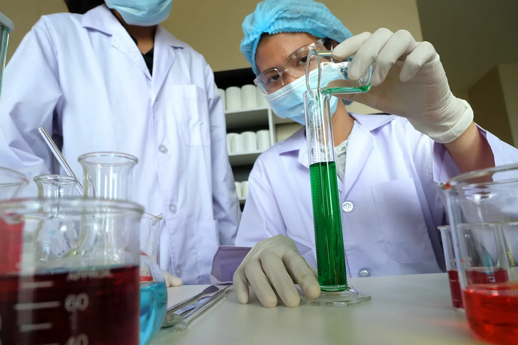 An analyst pouring a green solution into a graduated cylinder, beakers of coloured solutions alongside.