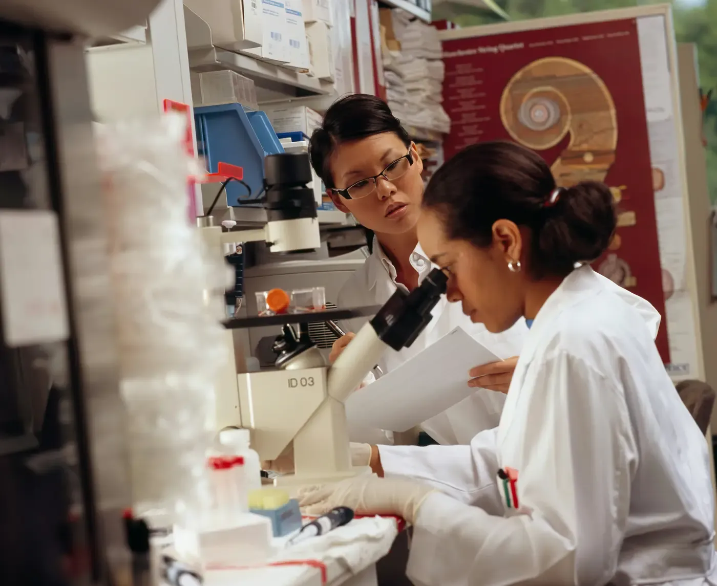 Two analysts at a microscope, one reading a printed result sheet.
