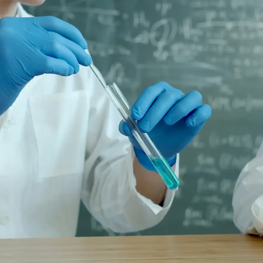 A gloved hand adding a drop from a pipette to a test tube, with equations chalked on the board behind.