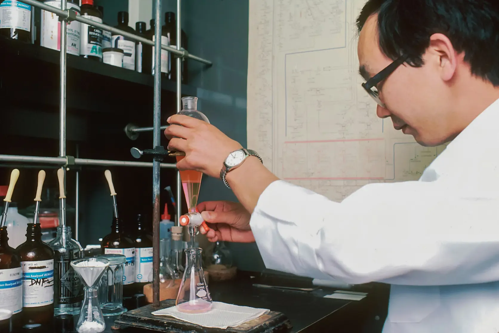 A chemist draining a separatory funnel into a flask, with reagent bottles and a synthesis scheme behind.